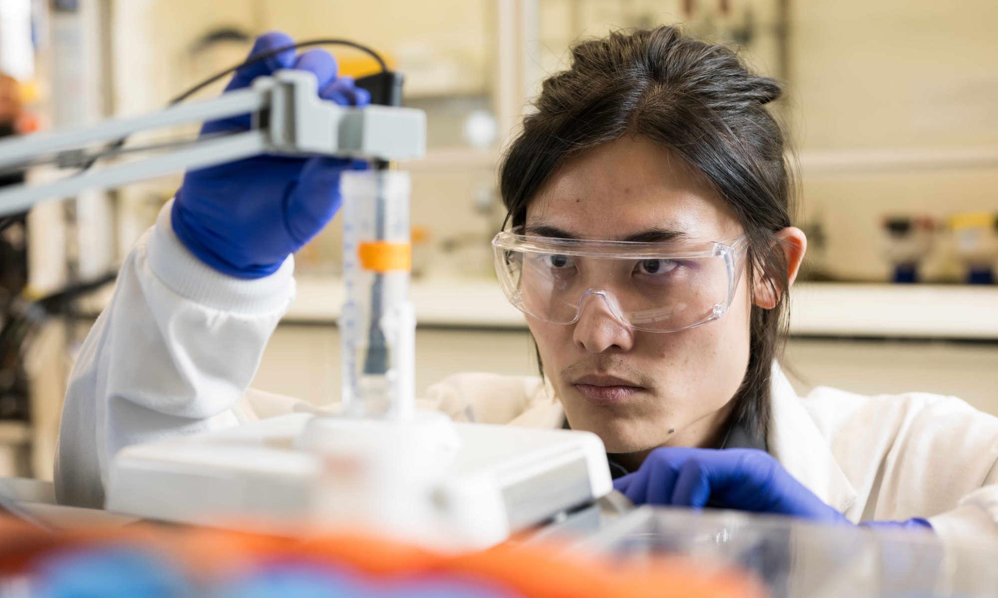 A scientist wearing lab googles and gloves examines a beaker of water containing PFAS chemicals.